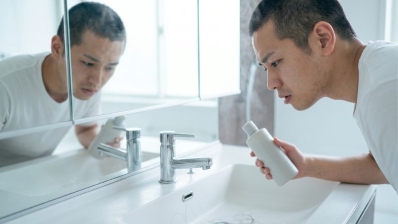 洗面台で抜け毛に気づき、薄毛の進行を不安に思う男性。AGA治療を開始する目安となる初期症状の不安。