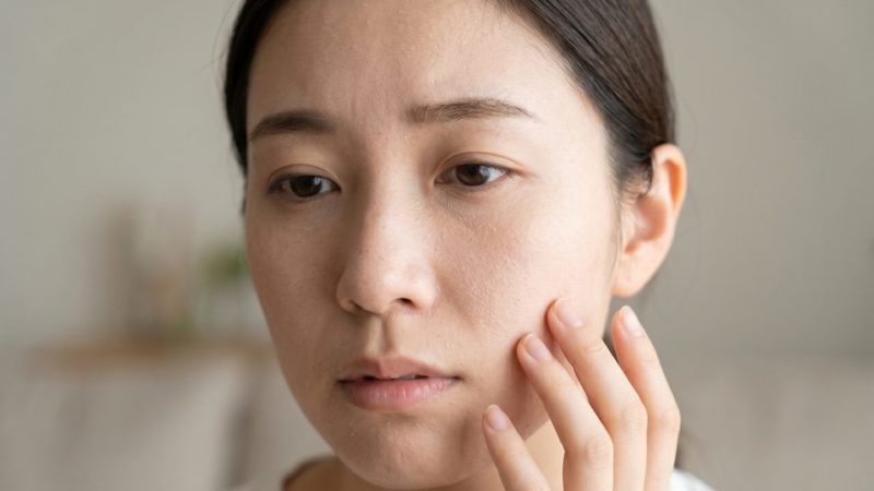 肌荒れや乾燥肌などの肌トラブルに悩み、鏡の前で頬に手を当てて状態を確認している女性。