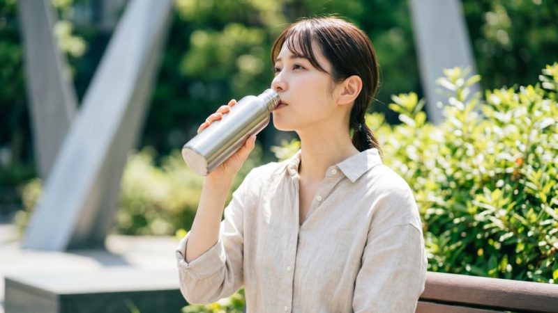 肌の潤いを保つためのインナーケアとして、屋外で水筒からこまめに水分補給を行う女性。