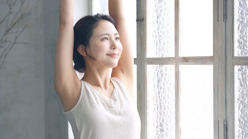 差し込む光を浴びながら、窓際でリラックスして深呼吸とストレッチをする女性。
