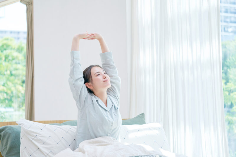 人気コラーゲンドリンクで健やかな毎日を。朝の光が差し込む寝室で、清々しく背伸びをするパジャマ姿の女性。