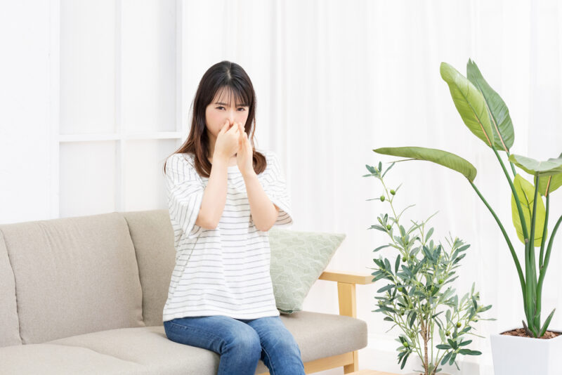 体臭が強くなる原因を解説。自分のニオイが気になり鼻をつまんで悩む女性と部屋の様子。
