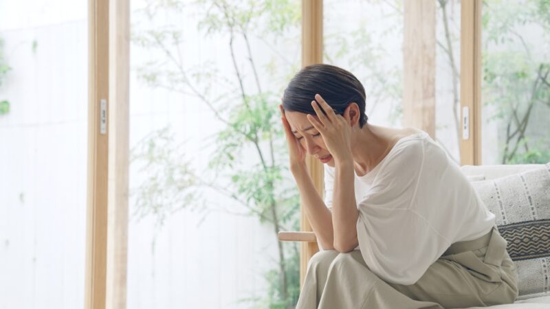 疲労臭の原因となる蓄積したストレス。窓際で頭を抱え、疲れ果てて座り込む女性の深刻な様子。
