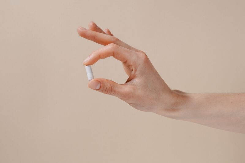 手軽に飲める尿酸値対策サプリメント。指先で摘んだ1粒の白いカプセル剤のアップ。