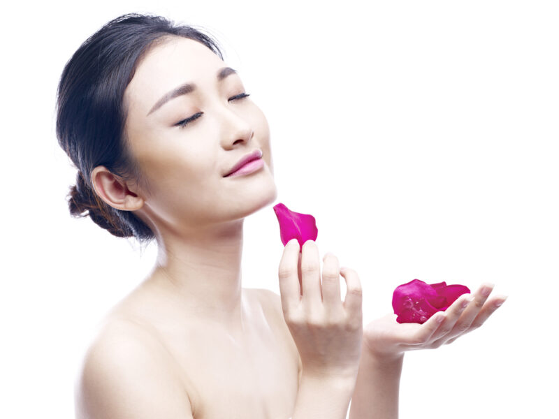 目を閉じて穏やかに微笑む女性。手のひらに乗せたピンク色のバラの花びらを、顎のあたりに優しく添えている。