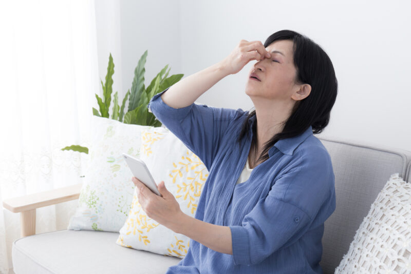 スマホ利用による目の疲れを感じ、目頭を押さえて顔をしかめるシニア世代の女性。