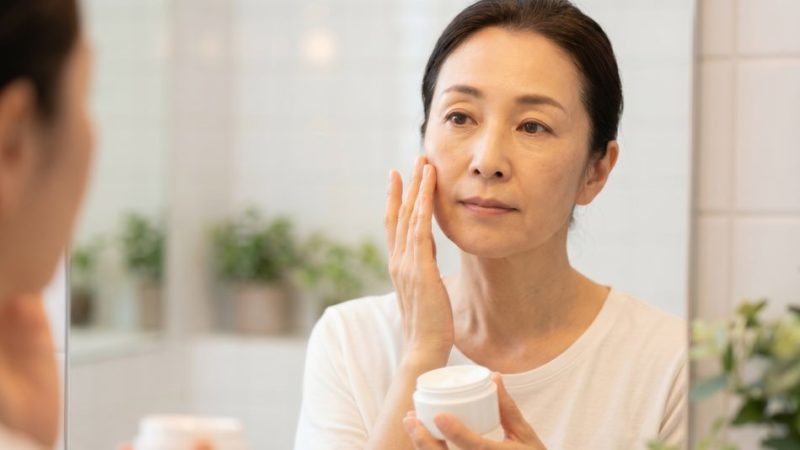 肌荒れや乾燥などの年齢によるサインを防ぐため、鏡の前で保湿クリームを頬に丁寧に塗り込む女性。
