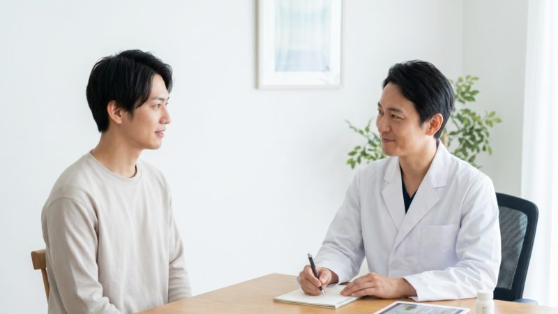 清潔感のある診察室で、白衣の医師と私服の男性が机を挟んで向き合い、男性がメモを取りながら話を聞いている。