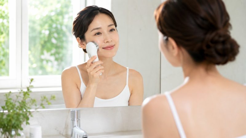 鏡の前で美顔器を使い、顔のたるみケアに励む女性。専門家が推奨する正しい使い方で、フェイスラインに沿って美顔器を滑らせる様子。
