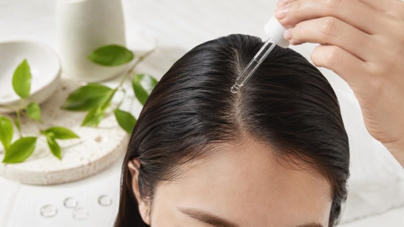 女性の頭頂部の分け目に、透明なスポイトから一滴のヘアケア液が垂らされる瞬間のクローズアップ。