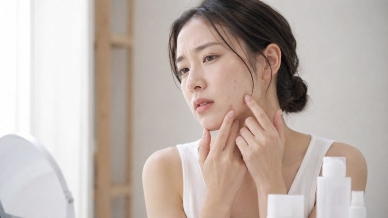 鏡を見ながら頬にできたニキビを指で直接触り、不安そうで険しい表情を浮かべている女性のクローズアップ。