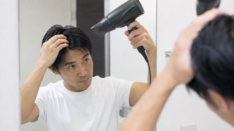 鏡に向かって立ち、左手で髪をかき上げながら、右手に持った黒いドライヤーの温風を髪の根元に当てて乾かしている男性。