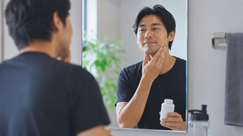 鏡の前で自身のコンディションをチェックしながら、ノコギリヤシサプリを手にする男性。リピート率の高いサプリによる毎日のケアを想起させる。
