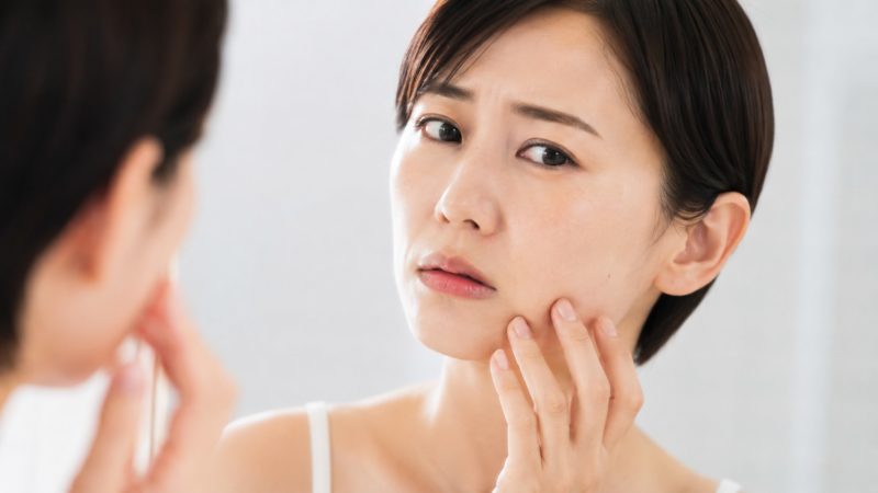 鏡を見ながら顔のたるみや肌のハリ不足を気にする女性。年齢とともに現れるフェイスラインの変化を確認し、本格的なたるみケアの必要性を感じている場面。
