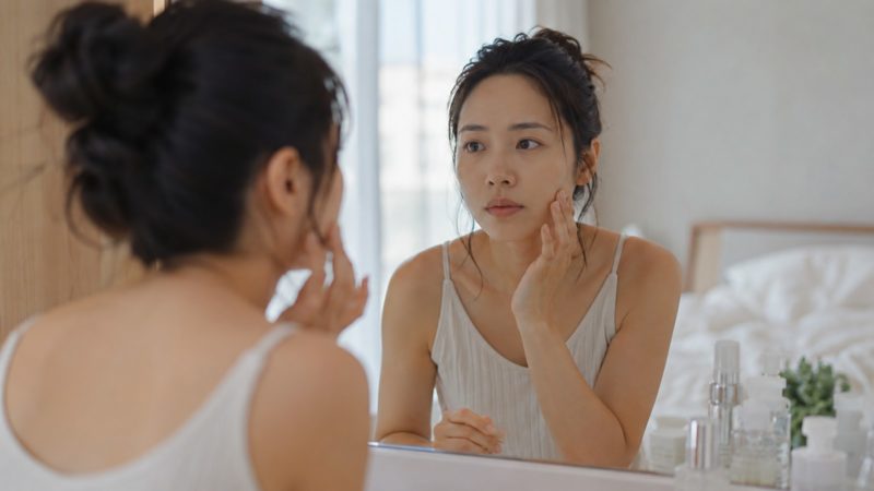 鏡に映る自分の顔を真剣な表情で見つめながら、指先で頬の感触を確かめ肌の状態を確認する女性の背後からの視点。