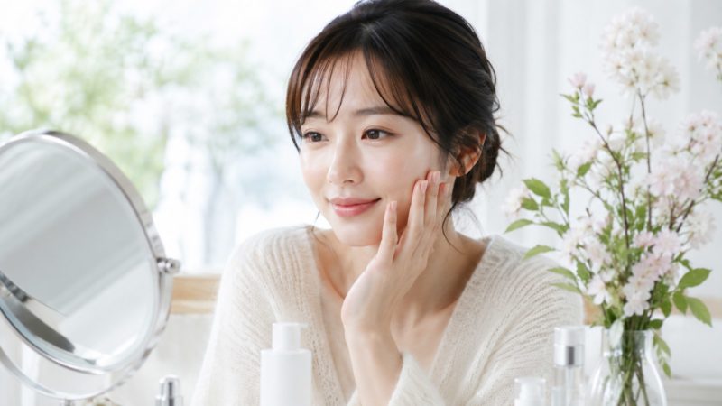 肌荒れサプリの効果を実感し、鏡の前で微笑む女性。傍らには白い花とスキンケア用品が置かれた明るいドレッサー。