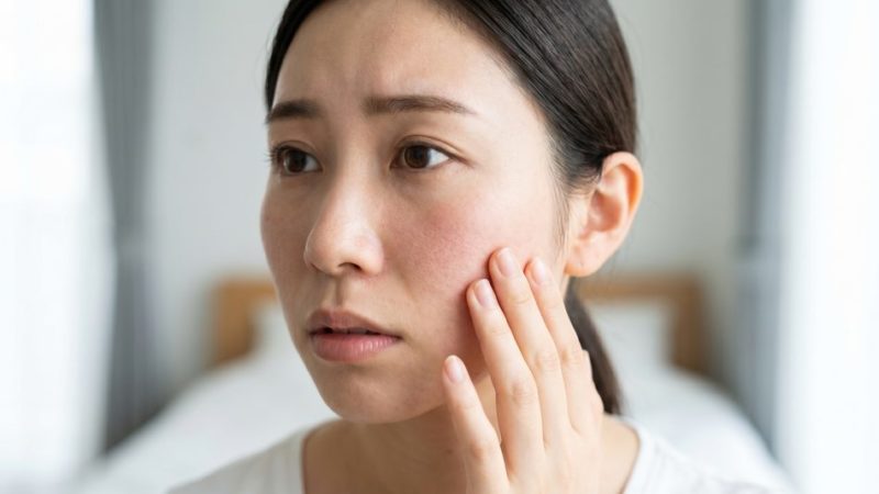 乾燥や赤みなどの肌荒れトラブルを気にして、不安そうな表情で自分の頬に触れる女性。