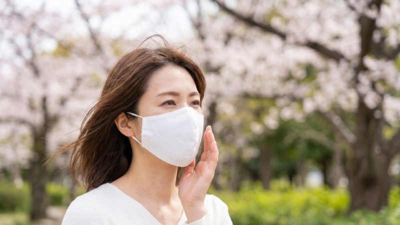 春の花粉や乾燥などの外部刺激による肌荒れを防ぐため、桜の咲く公園でマスクを着用して過ごす女性。